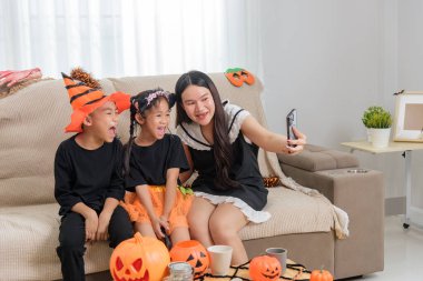 Kanepede kostüm giymiş genç bir kadın iki gülümseyen çocukla bir telefon fotoğrafı çekiyor. Ayrıca kostümlü, balkabaklı, kapalı alanda bayram teması sergiliyor..