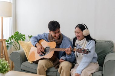 Kış, mutlu Asyalı çift, ev içinde rahat, insanlar müzik anından zevk alıyor, erkekler kadınlar için akustik gitar çalıyor, sıcak atmosfer, neşeli kış günü rahatlaması sağlıyor..