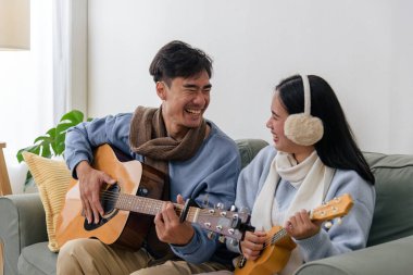 Winter brings couple happy people indoor home enjoying music, Asian man plays guitar, woman strums ukulele wearing ear muffs and scarf, cozy warm winter mood indoors.