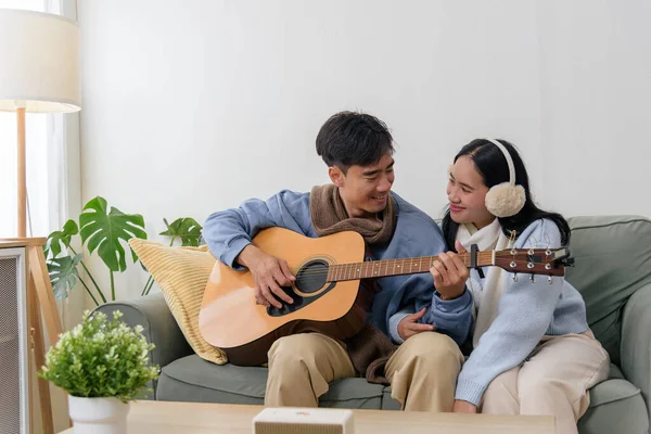 Winter day finds happy couple, young people building family, home offers warm indoor comfort. Man plays guitar, woman enjoys music, cozy winter moments shared, smiles exchanged.