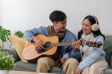 Winter day brings happy couple home for cozy indoor music session, people enjoy relaxed evening on couch, man playing guitar for woman in warm winter attire, creating joyful memories.