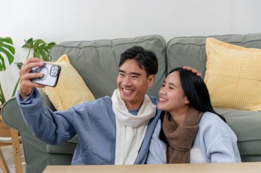 Winter day sees happy couple at home, people share cozy indoor moments, capturing joyful memories with smartphone selfie, embracing warm winter comfort and happiness.