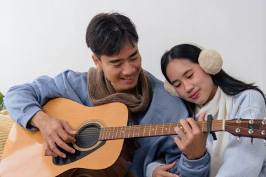 Winter day indoors, happy young couple enjoys cozy home bonding with music, man plays guitar, woman rests on shoulder, creating warm winter memories.