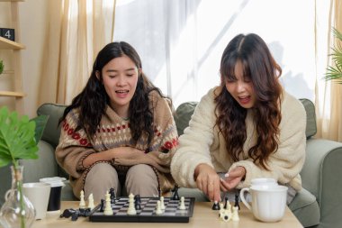 Winter day brings warm friendship, young women friends enjoy cozy home activities, playing chess indoors during cold winter, sharing laughter, happiness in comfortable space.