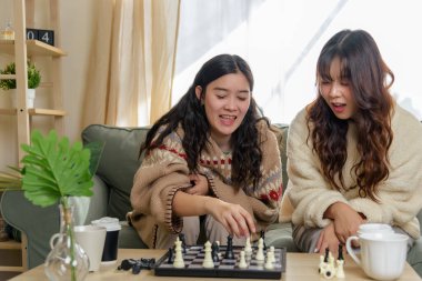 Winter day friendship brings young women friends together at home, enjoying warm chess game, cozy sweaters and hot drinks enhance pleasant indoor companionship, perfect winter memories at home.