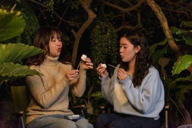 Winter night camp brings happy women friends outdoor in lush forest, people gather enjoying cozy evening winter marshmallow treats, blowing softly on sweet snack.