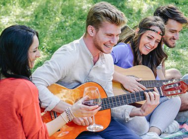 Gitar çalmaya ve kırmızı şarap bir Park açık - mutlu gençler eğleniyor birlikte bir piknik - dostluk, gençlik yaşam tarzı kavram yapma çimenlerin üzerinde oturan yudumlarken gülüyor arkadaş grubu