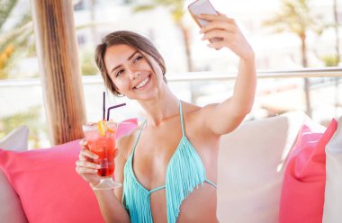 Bir beach club - çekici kız bir tropikal kokteyl ve deftere nakil fotoğraflar üzerinde sosyal onun networs onu tatil sırasında içme eğleniyor mobil akıllı telefon ile bir selfie alarak genç güzel kadın