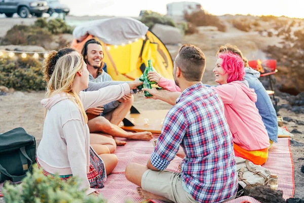 Happy friends toasting with beers at barbecue dinner on the beach - Young people camping with tent having fun cheering and drinking beer - Friendship and youth lifestyle vacation concept