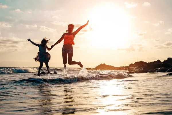 Happy loving family of mother and daughter jumping in the water at sunset on the beach - Mom with her kid having fun together on a summer day - Concept of parents summer vacations