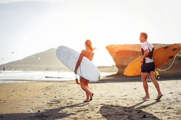 Surfers couple running together with surfboards on the beach at sunset - Sporty friends having fun going to surf - Travel, vacation, sport lifestyle concept