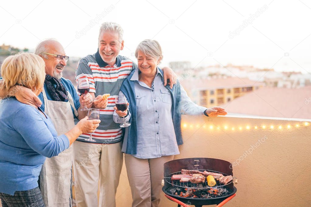 Happy senior friends having fun drinking red wine at barbecue dinner in terrace - Mature people dining and laughing together on rooftop - Friendship and elderly lifestyle concept