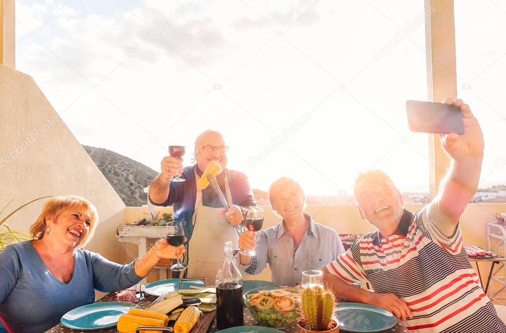Happy seniors taking a selfie with mobile phone and making a barbecue on the rooftop - Retired people having fun eating and drinking red wine laughing together - Cheerful elderly activity concept