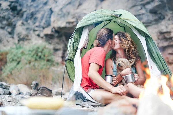 Travel couple camping in rock mountains after a trekking day - Happy people drinking hot tea inside tent with their dog next to bonfire - Sport, relationship, love and vacation concept