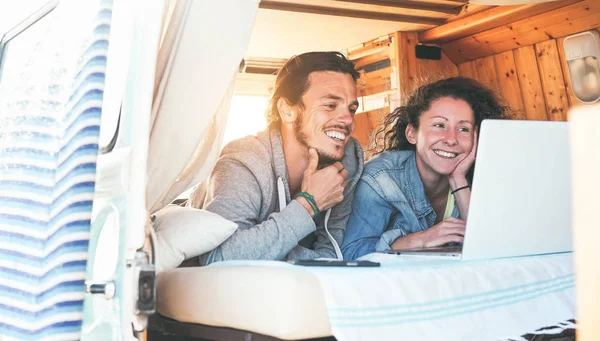 Happy couple watching videos on their computer inside minivan at sunset - Travel couple using laptop during their journey on a vintage mini van with wood interior - Vacation, love, technology concepts