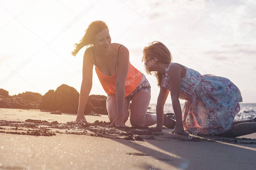 Mother and daughter having fun drawing with sand at sunset on tropical beach - Happy mom playing with her kid in vacation - Love, happiness and family lifestyle concept