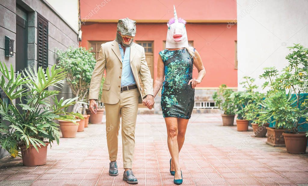 Crazy senior couple wearing dinosaur and unicorn mask - Mature trendy people having fun masked at carnival parade - Absurd, eccentric, surreal, fest and funny masquerade concept