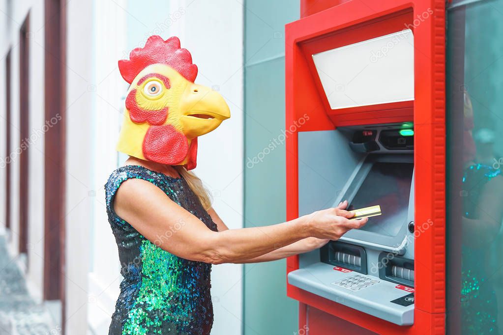 Mature woman wearing cock mask withdraw money from bank cash machine with debit card - Surreal image of half human and animal - Absurd and crazy concept of ATM advertise