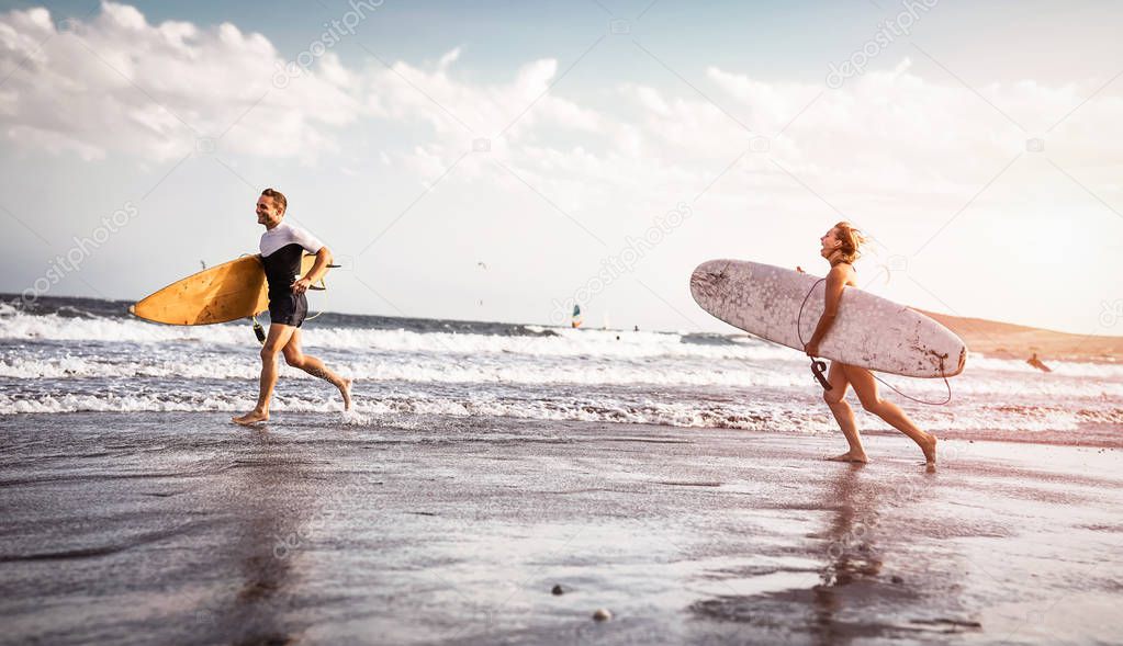 Happy surfers running with surfboards along the sea shore - Sporty couple having fun going to surf together at sunset - Extreme surfing sport, sportive people and youth relationship lifestyle concept