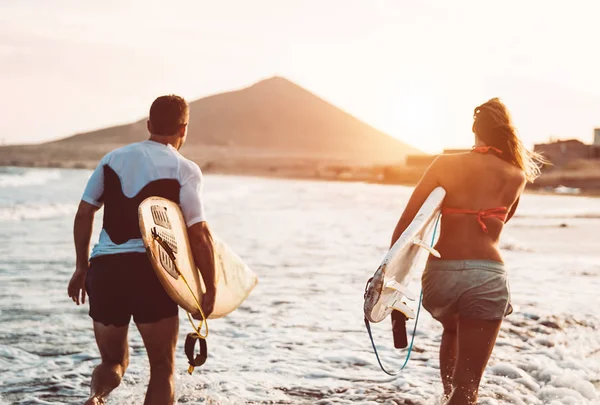 Happy surfer couple running with surfboards along the sea shore - Sporty people having fun going to surf together at sunset - Extreme surfing sport and youth relationship lifestyle concept — Stock Photo, Image