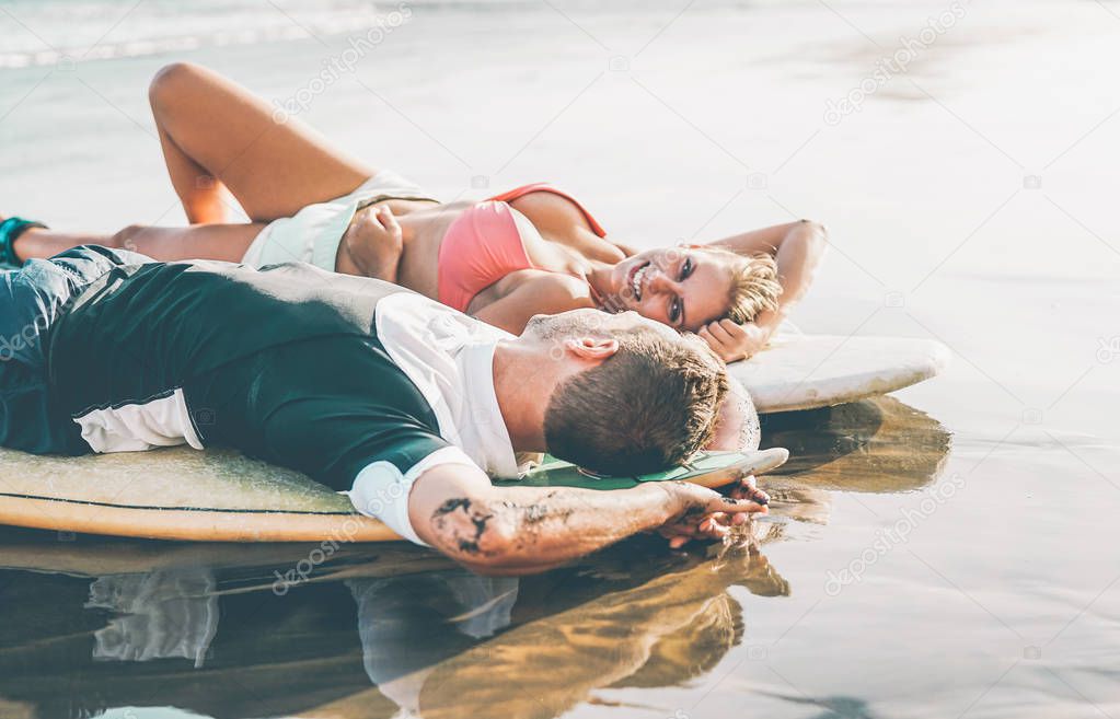 Happy couple of surfers lying on surfboards on the beach after a sporty day - Healthy lovers laughing looking each others relaxing on surf longboards - People, sport and love relationship concept