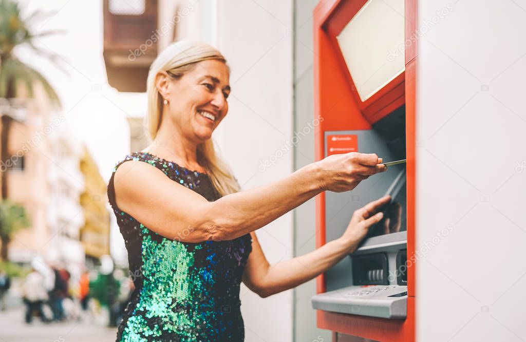 Happy mature woman withdraw money from bank cash machine with debit card - Senior female doing payment with credit card in ATM - Concept of business, banking account and lifestyle people