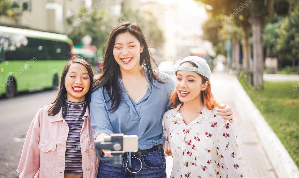 Happy Asian friends vlogging in city bus station - Trendy young people using gimbal smartphone outdoor - Friendship, technology, youth lifestyle and social media concept