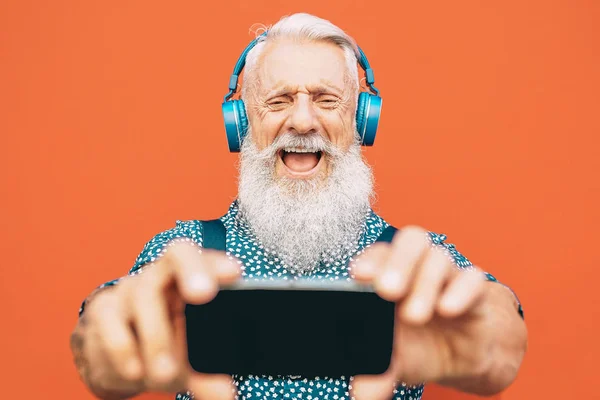 Senior skäggiga man göra Selfie med mobiltelefon medan du lyssnar på hans favoritspellista med hörlurar-mode mognat manliga ha kul med smartphone Apps utomhus-äldre, teknikkoncept Stockfoto