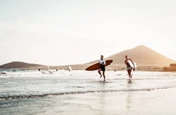 Happy surfer couple running with surfboards along the sea shore - Sporty people having fun going to surf together at sunset - Extreme surfing sport and youth relationship lifestyle concept — Stock Photo, Image