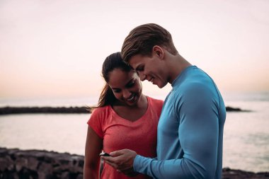 Genç çiftler ara veriyor ve açık havada cep telefonu kullanıyor. Sportif insanlar cep telefonu teknolojisi uygulamalarıyla eğleniyor. Sağlıklı, teknoloji ve gençlik yaşam tarzı kavramı.