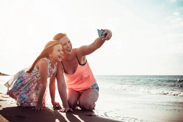 Madre e hija tomando selfie con teléfono inteligente móvil mientras juegan en la playa tropical al atardecer - Familia feliz divirtiéndose con nuevas aplicaciones de tecnología para las redes sociales - Concepto de paternidad —  Fotos de Stock