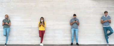 Cerrahi maske takan gençler cep telefonlarını kullanarak sosyal mesafeyi koruyorlar - Milenyum arkadaşları Corona virüsü salgını sırasında akıllı telefonlardan medya trendlerini izliyorlar - Gençlik ve teknoloji kavramı