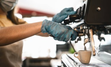 Corona virüsünün yayılmasını önlemek için cerrahi maske ve eldiven takarak kahve yapan genç kadın yan görüşü - Bar sahibi güvenliği çalışıyor - Sıcak içecekler ve covid-19 kural kavramı