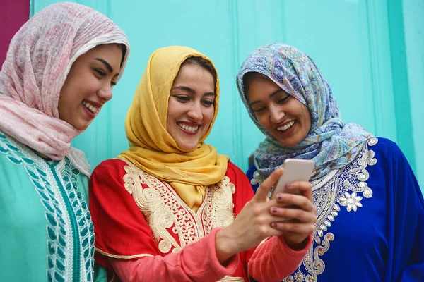 Young Muslim friends watching on mobile phone outdoor - Happy Arabian girls addicted to new technology smartphone app for social media - Youth millennial people and religion concept