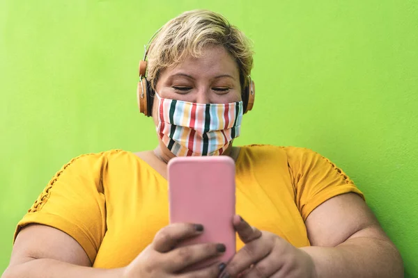 携帯電話を使用し コロナウイルス感染を避けるために顔保護マスクを着用しながら ワイヤレスヘッドフォンで音楽を聴くプラスサイズの女性 多様な人々と技術の概念 — ストック写真