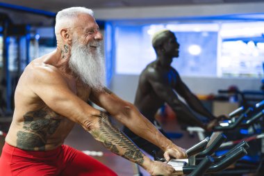 Spor salonunda bisiklet egzersizi yapan yaşlı ve genç erkekler - sağlık merkezinde bisiklet makinesi üzerinde farklı yaş ve ırk eğitimi alanlar - spor aktivitesi ve fitness yaşam tarzı konsepti