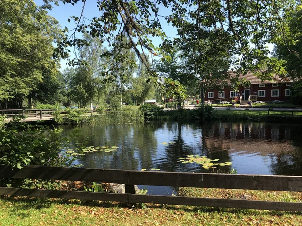 Cabana vermelha / casa e vegetação por um pequeno riacho / lagoa com água caindo lentamente. Cores e luzes de verão agradáveis. Hogbo Bruk, Suécia, julho de 2019 . Fotos De Bancos De Imagens