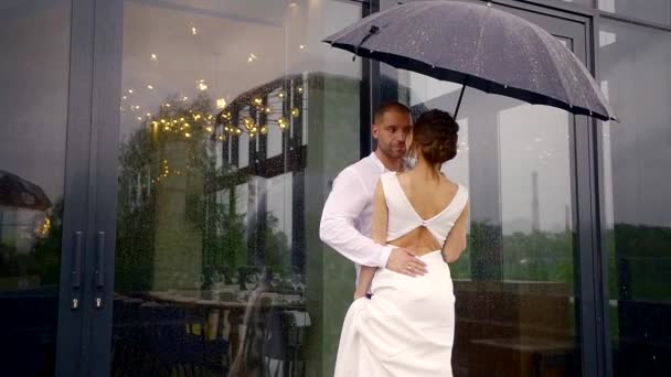 a man and his beautiful bride dressed in a white wedding dress are standing together under an umbrella, its raining outside, and the newlyweds are hugging and talking to each other