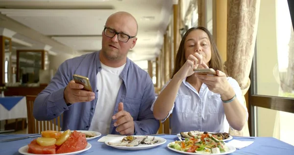 adult spouses are dining in cafe and using smartphones, looking on displays and chatting