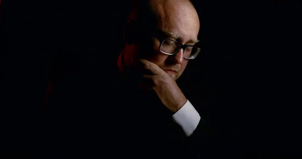 Portrait of a bald sad man in glasses and a business suit with a tie, it is on a black background. He charges the gun and puts it in his temple.