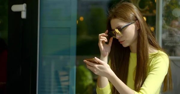 Dark-haired girl with long straight hair sitting in a cafe on the street in dark reflective glasses. She is holding a smartphone and is chatting with someone, smiling.