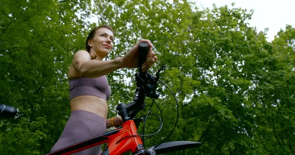 Beautiful slim girl in a t-shirt and the tights carries the bike by the handlebars and come along among the trees and green grass.