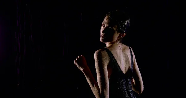 Beautiful wet girl in black tight dress dancing on a black background in heavy rain.