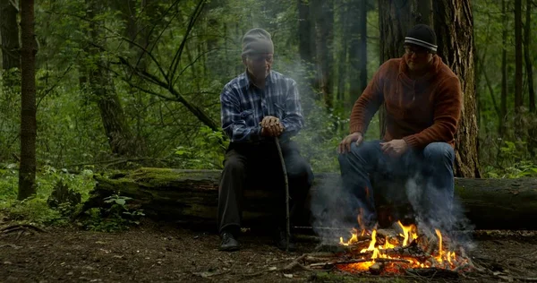 guy in brown hoodie sits near aged man with stick in forest