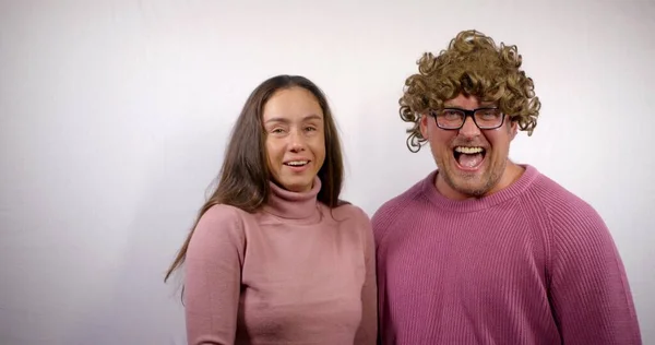 A positive middle-aged couple, a man in a curly wig and a dark-haired woman dressed in pink sweaters, stand in the Studio on a white background, blowing their mouths, smiling, joking, laughing.