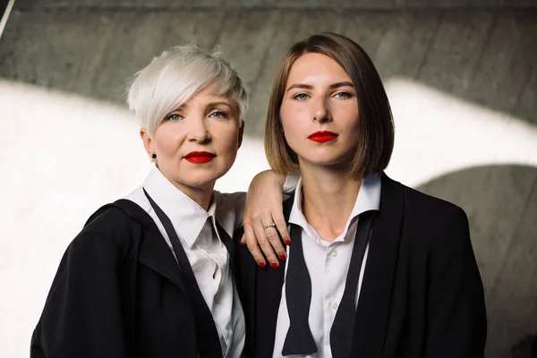portrait of two women in classic suits and red lips  posing against geometrical concrete wall outdoors