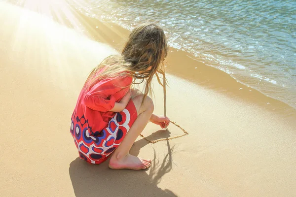 Счастливый ребенок рисует на песке на фоне моря — стоковое фото