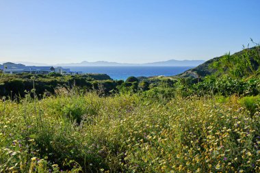 ön planda yeşil bir alan ve arka planda dağlar ile deniz görünümü, sol tarafta evler ile