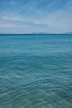 turkuaz renk sakin deniz ve mavi gökyüzünde bazı bulutlar, yelkenliler ve dağlar çok uzak arka planda