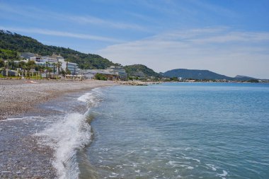 otel ve arka planda bazı küçük dağlar, mavi gökyüzü ve berrak su ile Akdeniz'de plaj